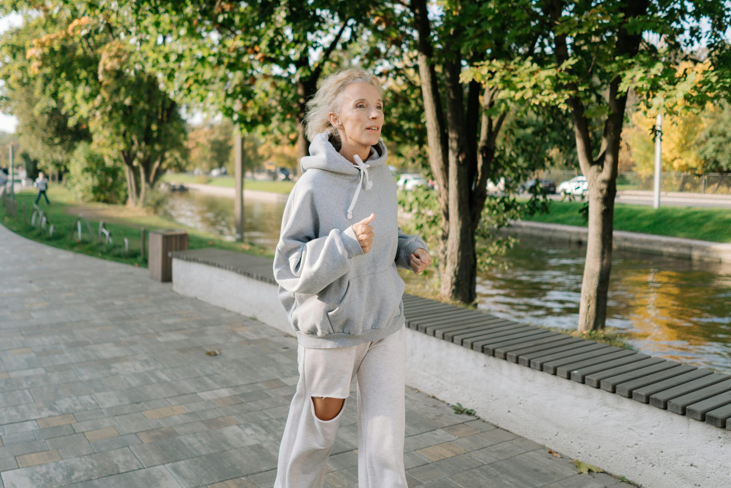 Senior woman in gray hoodie jogging along a park pathway by a canal, embracing an active lifestyle.