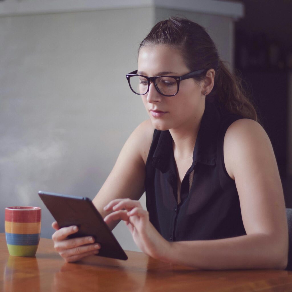 Caucasian woman in glasses using a tablet at home, enjoying coffee, looking engaged.