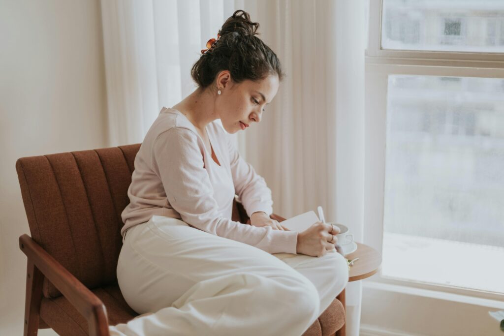 A woman comfortably journaling by a window, surrounded by soft natural light, in a cozy indoor setting.